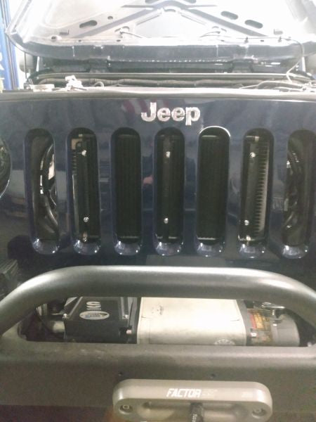 Wicked Wrench Motorsports dual transmission and power steering cooler bracket view looking directly through the vertical factory grille slots of a Jeep Wrangler JK